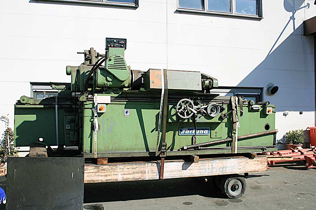 Grüne Rundschleifmaschine von Fortuna, auf Kanthölzern verzurrt auf einem Rollwagen vor einem Gebäude, mit Schleifspindelstock und Werkstückspindelstock entlang des Betts